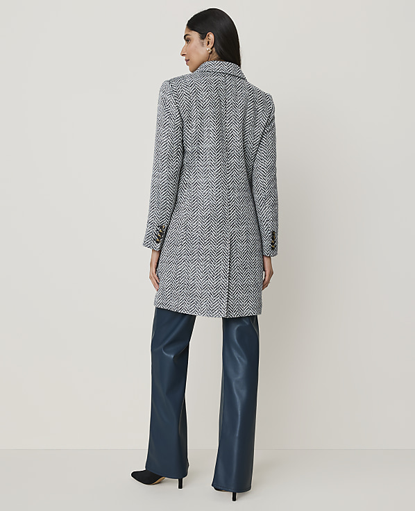 A woman in a patterned coat and teal pants stands against a plain, light-colored background.