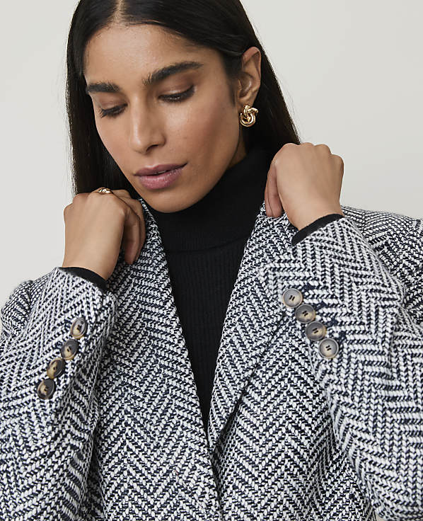 A woman in a herringbone blazer and black turtleneck looks down against a plain background.