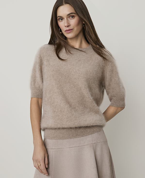 A woman in a fuzzy beige sweater and skirt stands against a plain, light background.