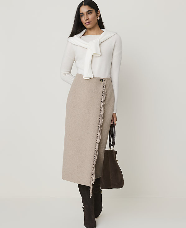 A woman in a cream sweater and beige skirt stands against a plain, light background, holding a dark brown bag.