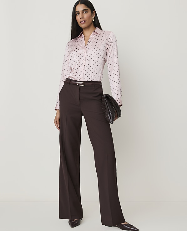 A woman in a pink polka dot blouse and brown wide-leg pants stands against a plain white background, holding a woven clutch.