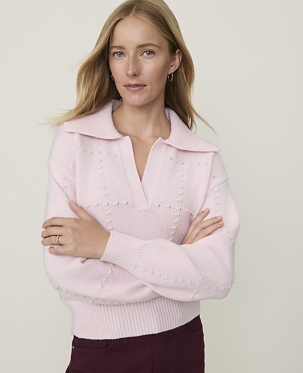 A woman in a pink collared sweater stands with her arms crossed against a plain background.