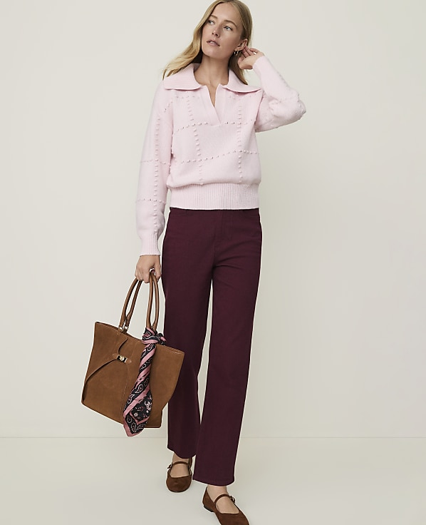 A woman in a pink sweater and burgundy pants stands against a plain background, holding a brown tote bag.