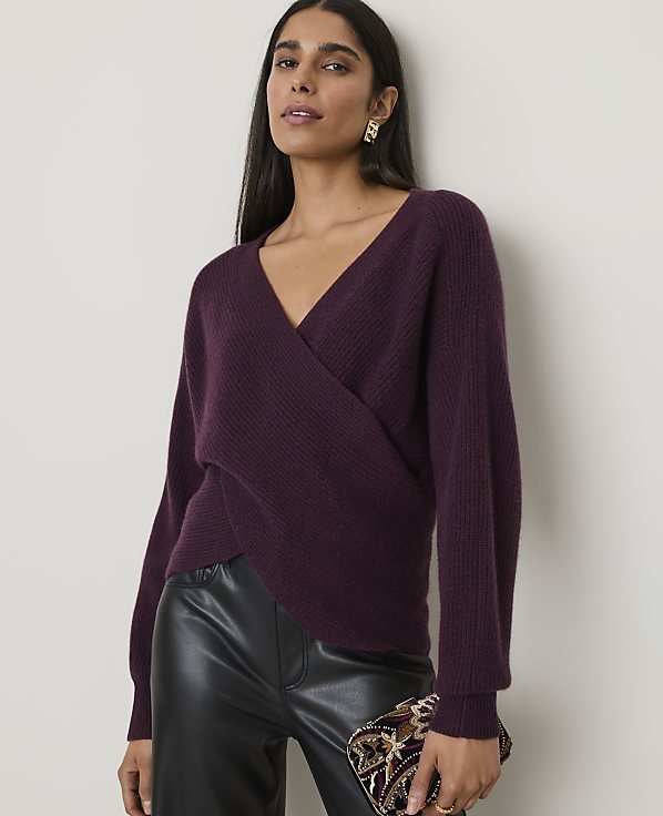 A woman in a plum-colored wrap sweater and black leather pants holds a jeweled clutch in front of a plain, light-colored wall.