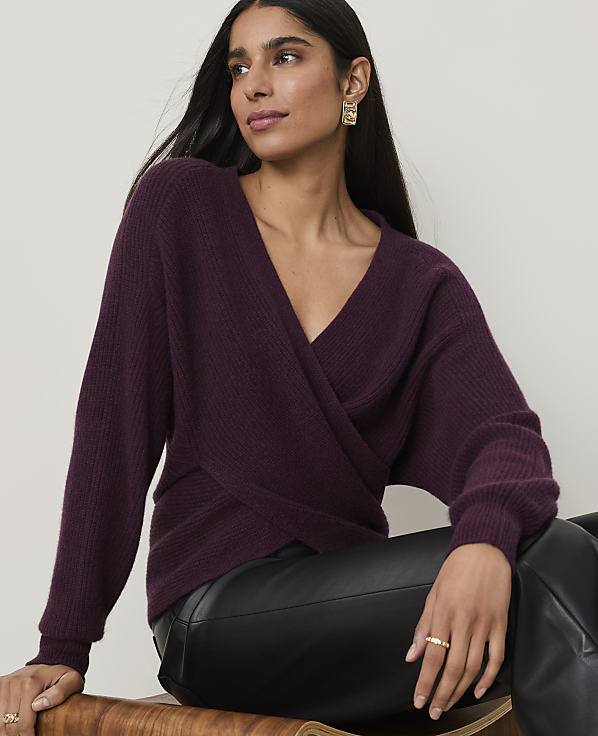 A woman in a plum-colored wrap sweater and black leather pants sits against a plain, light background.