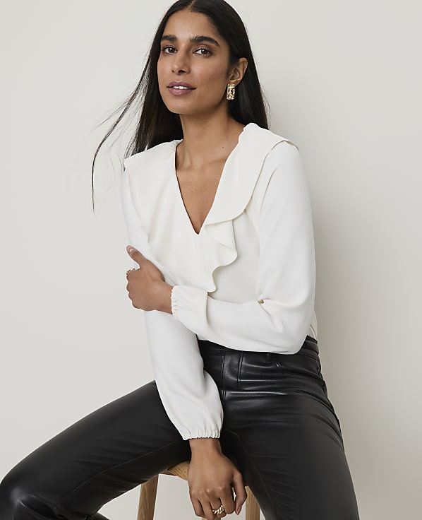 A woman in a cream ruffled blouse and black leather pants poses against a plain, light-colored background.
