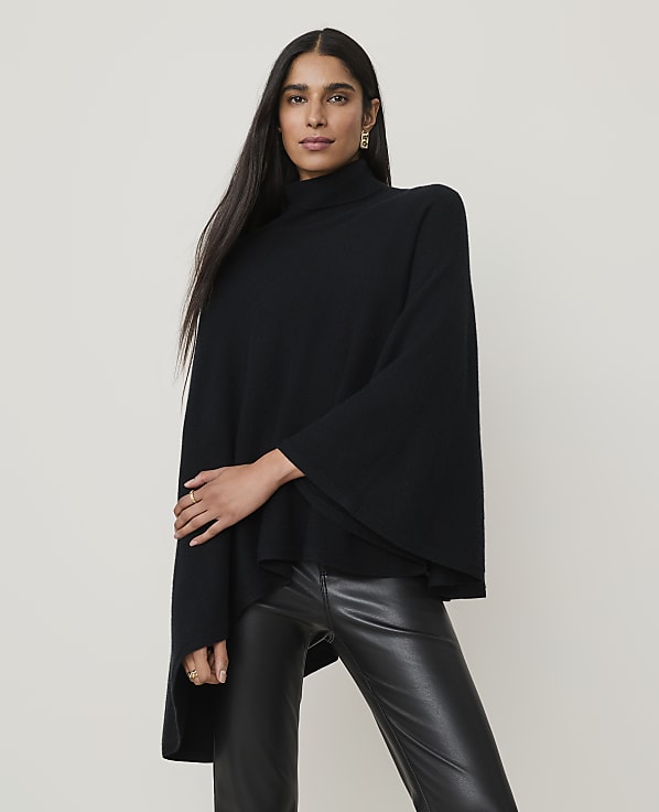A woman in a black turtleneck poncho and leather pants stands against a plain, light-colored background.