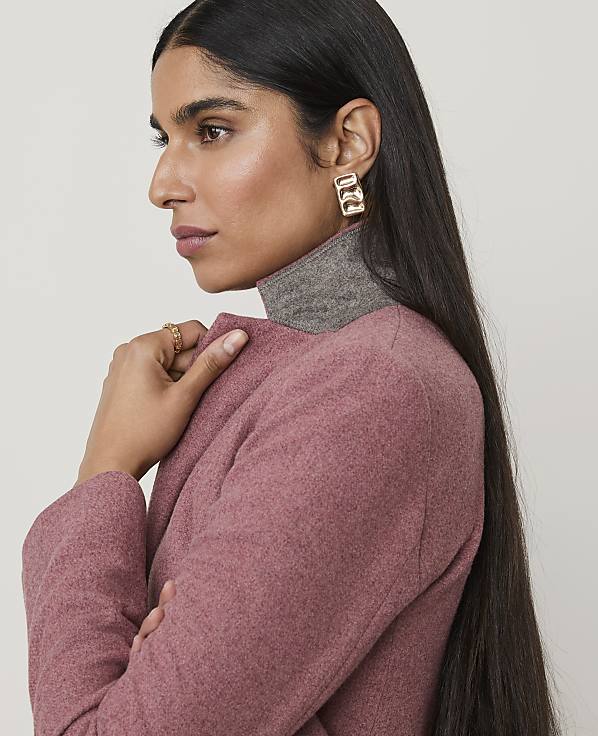 A woman with long dark hair and large gold earrings, wearing a pink coat over a grey turtleneck, is shown against a plain white background.