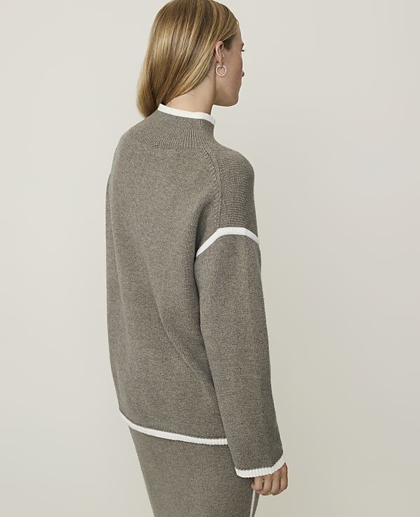 A woman in a taupe sweater and skirt stands against a plain, light gray background.