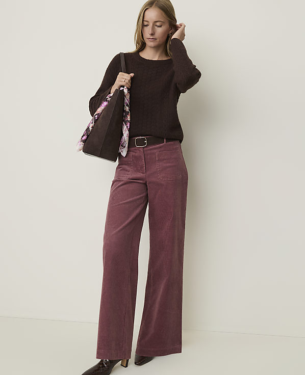 A woman in a brown sweater and pink corduroy pants stands against a plain, light-colored background, holding a tote bag.