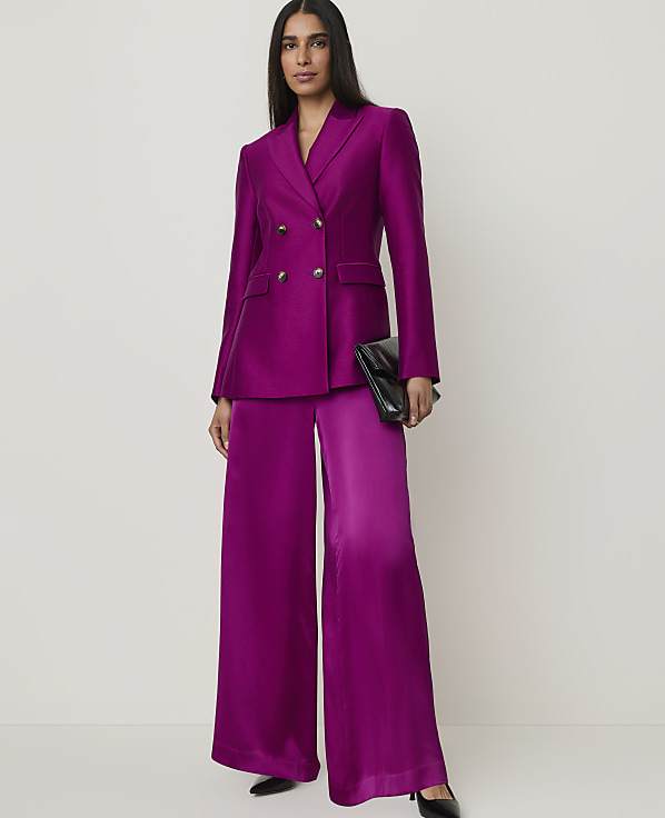 A woman in a vibrant magenta suit and black heels stands against a plain, light background, holding a black clutch.