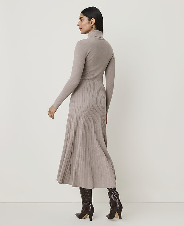 A woman in a beige turtleneck dress and brown boots stands against a plain, light-colored background.