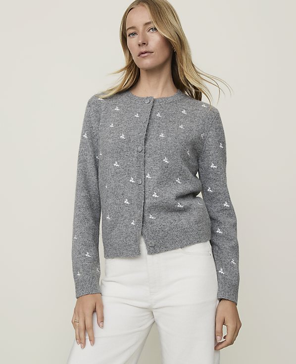 A woman in a grey cardigan with a white embroidered pattern stands against a plain grey background.