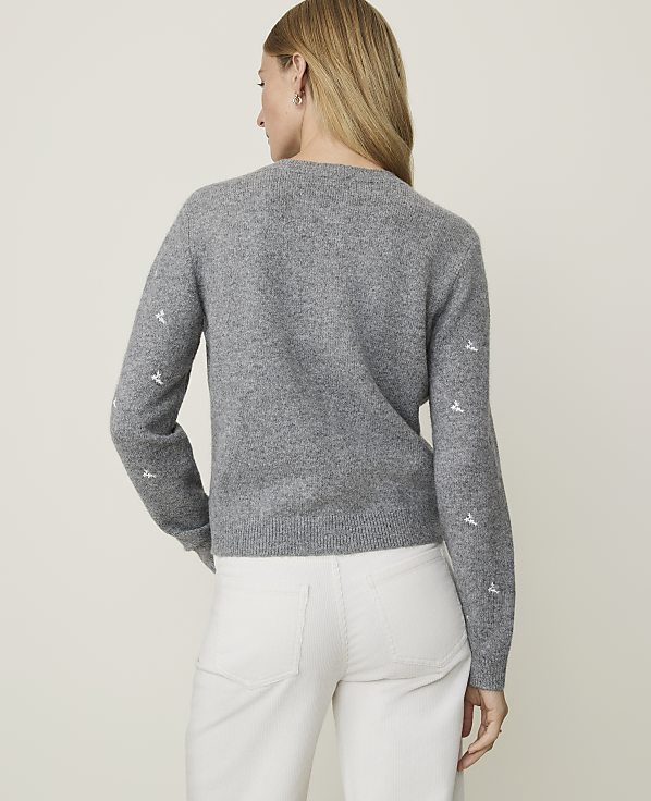 A woman in a grey sweater with embroidered details on the sleeves and white pants stands against a plain, light grey background.