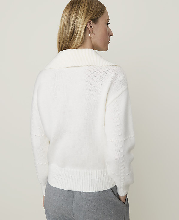 A woman in a white sweater and gray pants stands against a plain, light background.