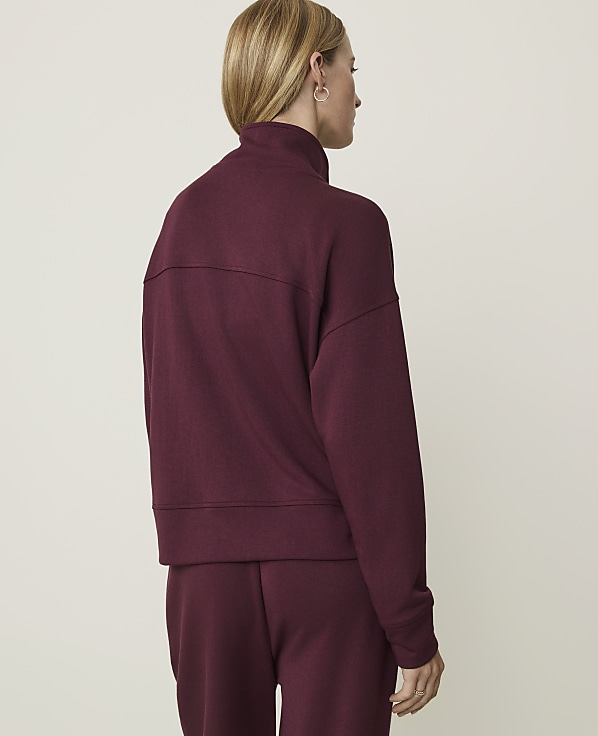 A woman in a burgundy sweatshirt and matching pants stands against a plain white background.