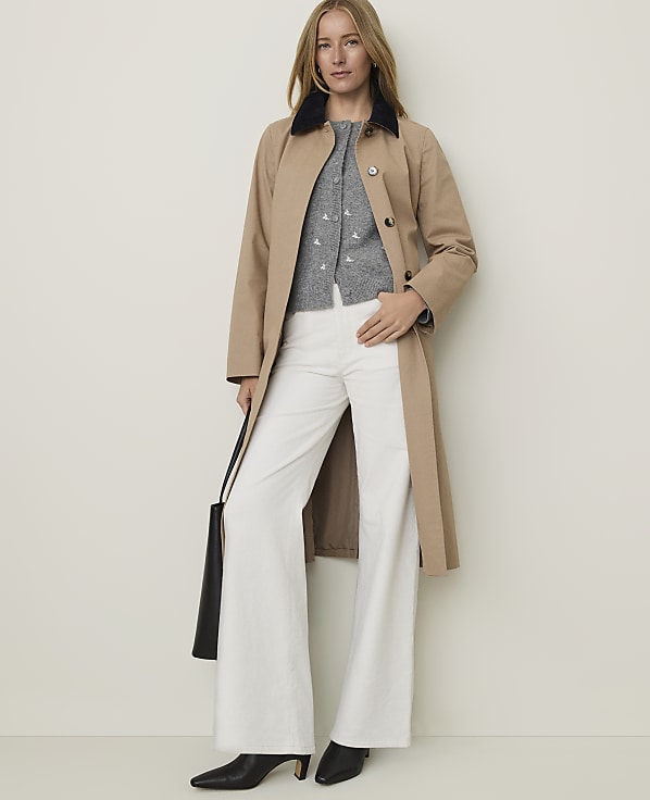 A woman in a tan trench coat, gray cardigan, and white flared pants stands against a plain, light-colored background, holding a black tote bag.