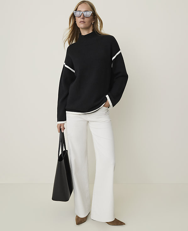 A woman in a black sweater and white pants stands with a black tote bag in front of a plain, light-colored background.