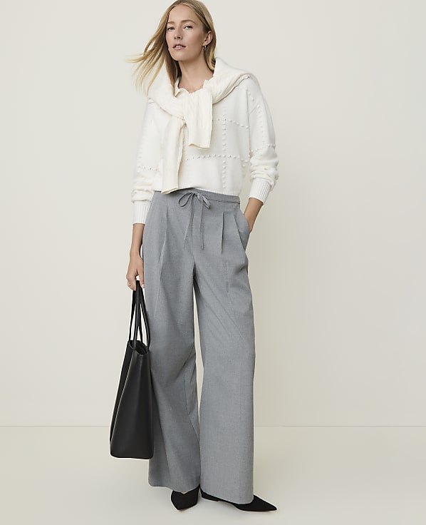 A woman in a white sweater and gray wide-leg pants stands with a black tote bag against a plain, light background.