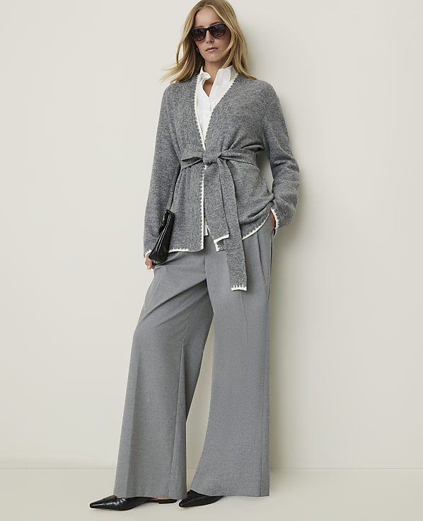 A woman in a grey belted cardigan and wide-leg pants stands against a plain, light-colored wall.