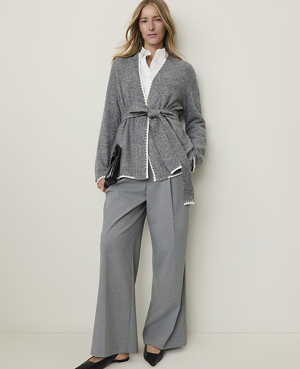 A woman in a gray cardigan and wide-leg pants stands against a plain, light-colored background.