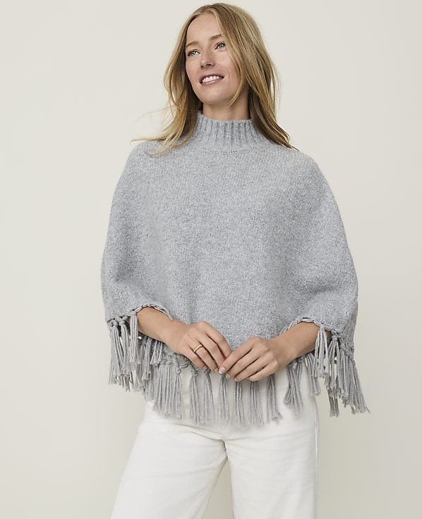 A blonde woman in a grey fringed poncho and white pants smiles against a plain, light grey background.