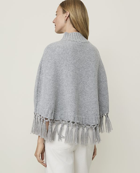 A woman in a grey poncho with fringe stands against a plain grey background.