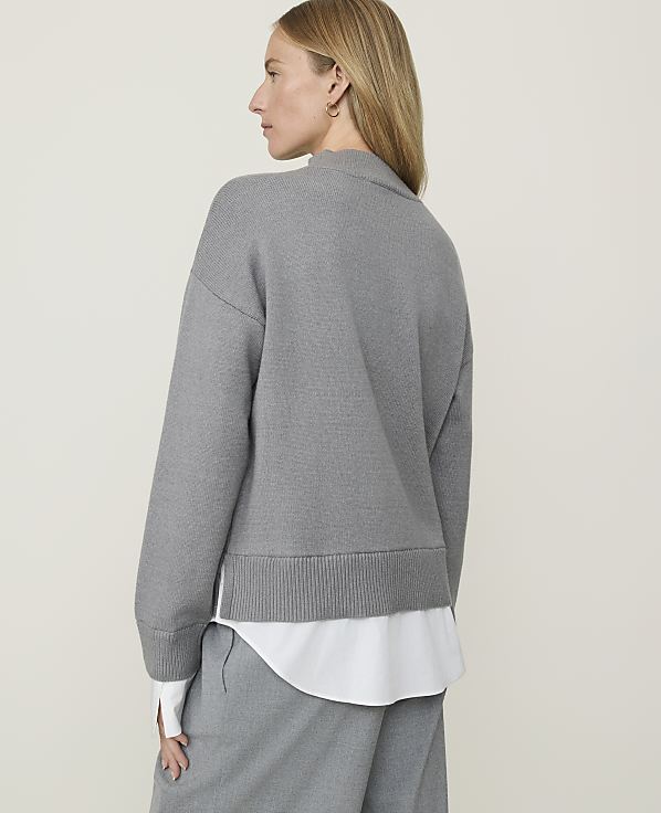 A woman in a grey sweater and white shirt stands against a plain, light grey background.