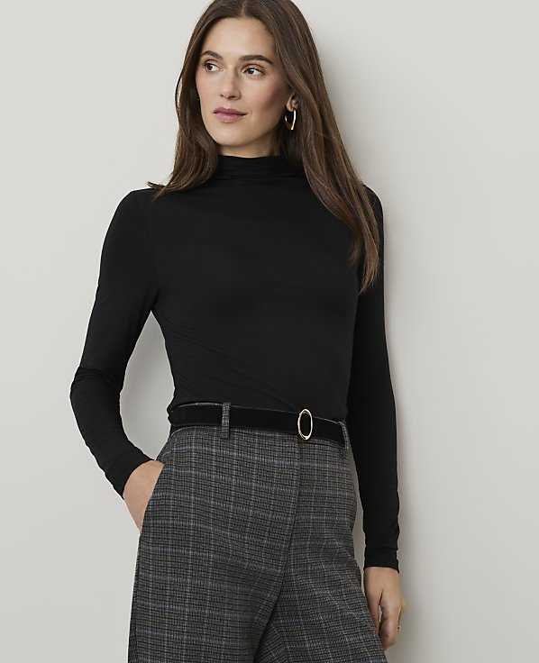 A woman in a black turtleneck and plaid pants stands against a plain, light-colored background.