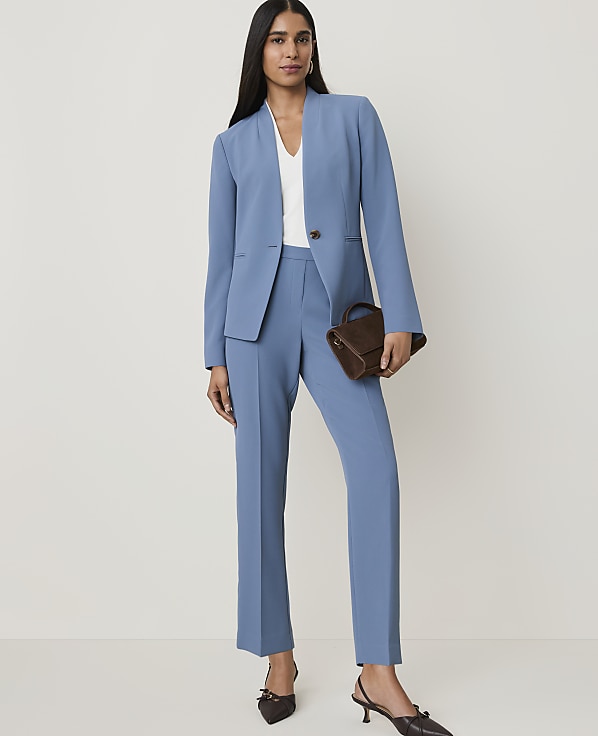 A woman in a blue suit sits on a stool, holding a woven bag, against a plain white background.