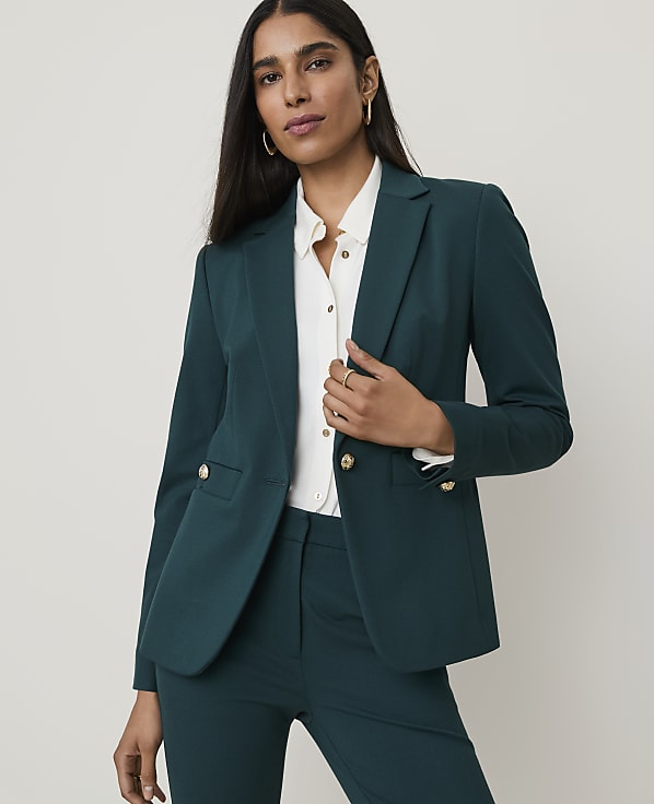 A woman in a teal suit stands against a plain, light background, holding a brown handbag.