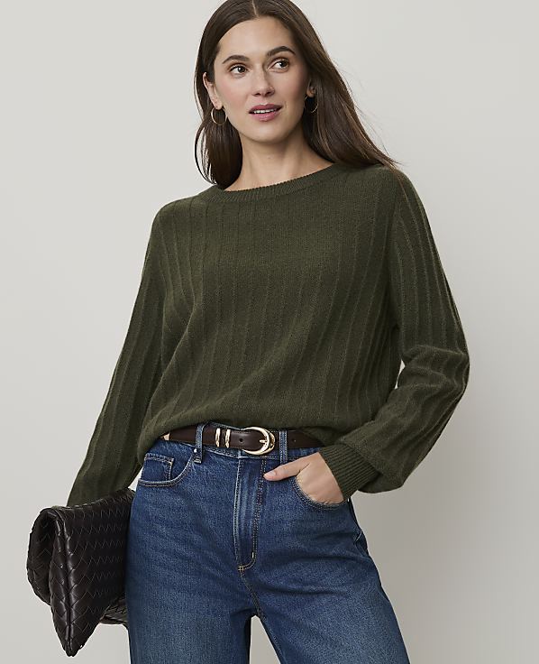 A woman in a green ribbed sweater and blue jeans holds a dark clutch in front of a plain background.