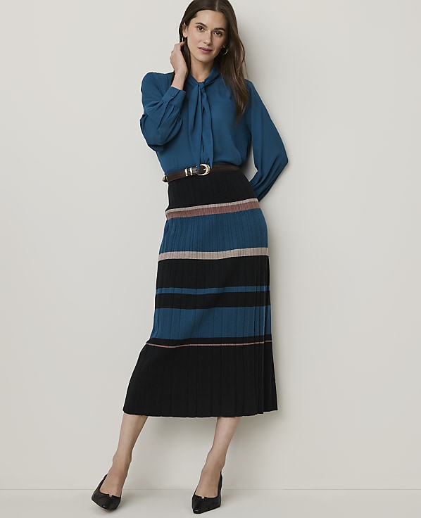 A woman in a teal blouse and a striped pleated skirt stands against a plain white background.