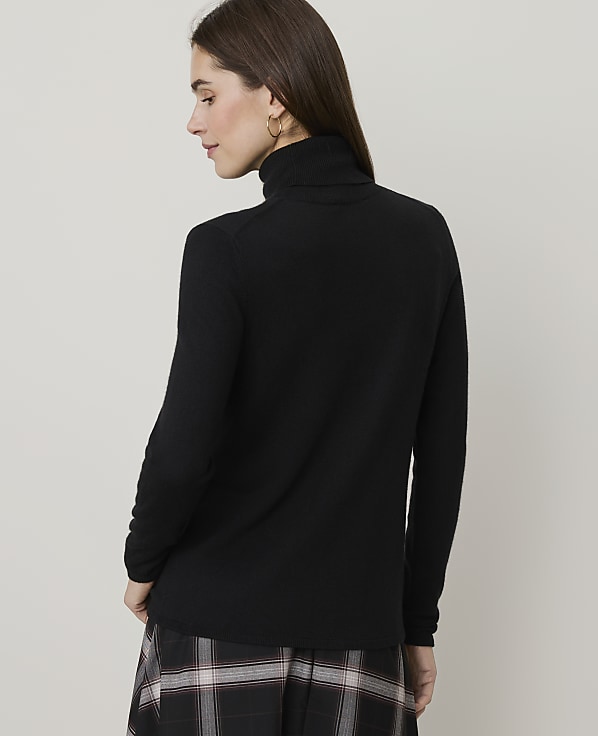 A woman in a black turtleneck sweater and plaid skirt stands against a plain, light gray background.