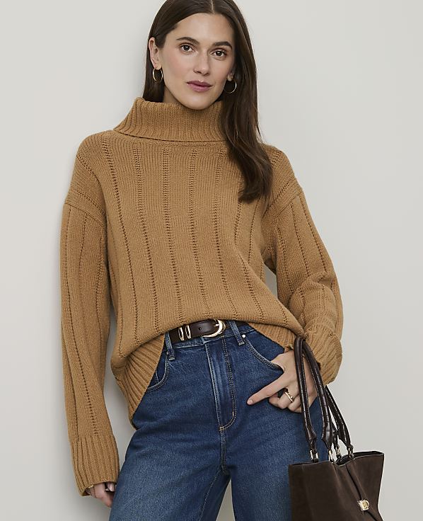 A woman in a camel turtleneck sweater and blue jeans stands against a plain background, holding a brown handbag.