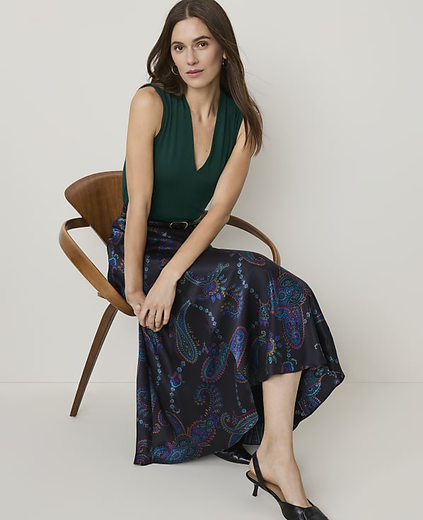 A woman in a green top and a patterned skirt sits on a wooden chair against a plain background.