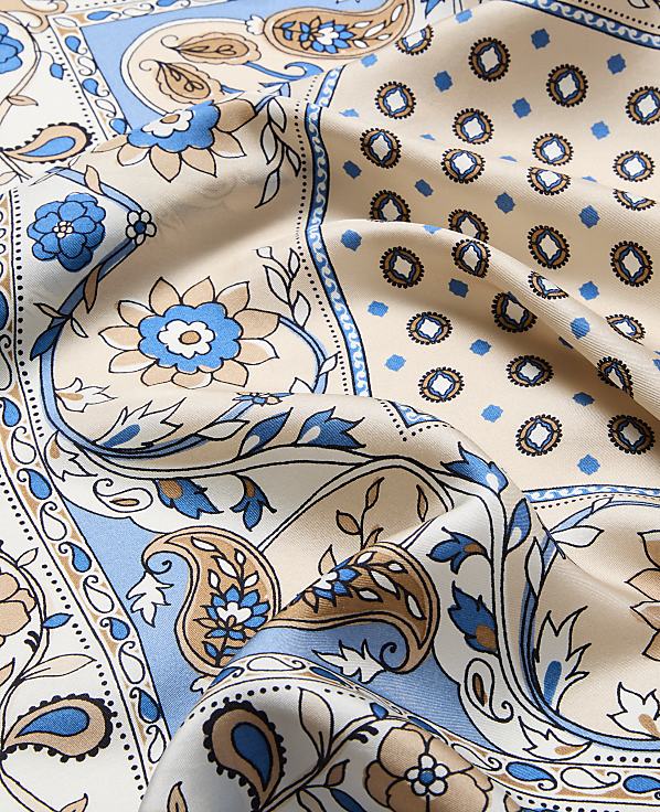 A close-up of a beige silk scarf with a blue and brown paisley and floral pattern in the foreground, set against a background of similar patterned fabric.
