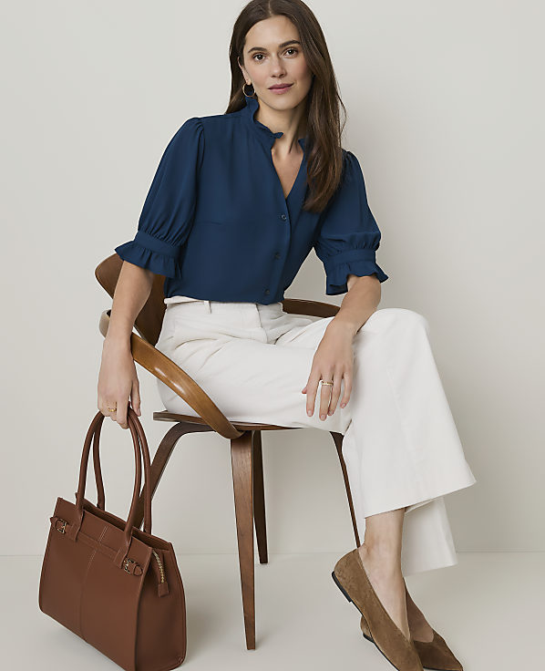 A woman in a teal blouse and white pants sits on a wooden chair holding a brown tote bag, with a plain, light-colored wall behind her.