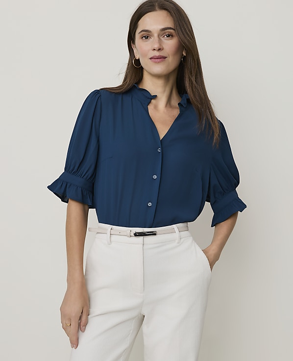 A woman in a teal blouse and cream pants stands against a plain white background.
