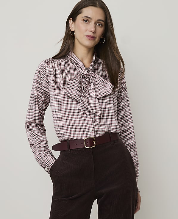 A woman in a pink plaid bow-neck blouse and brown corduroy pants stands against a plain gray background.