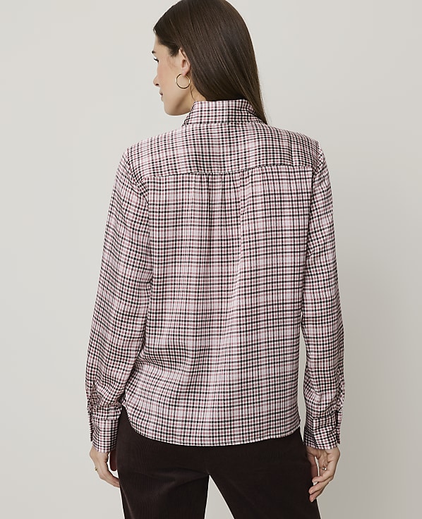 A woman wearing a pink plaid shirt and brown pants stands against a plain, light-colored background.