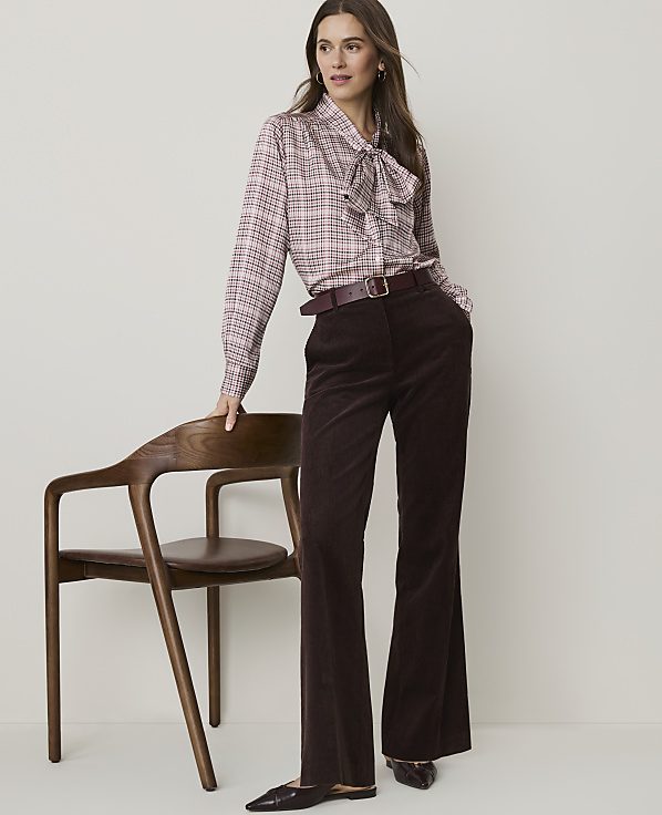 A woman in a pink plaid blouse and brown corduroy pants leans against a wooden chair in front of a plain background.