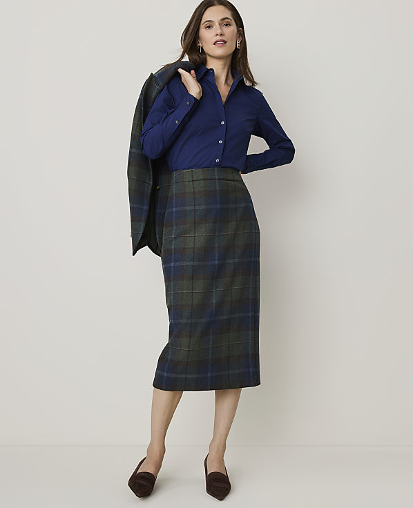 A woman in a blue button-down shirt and a plaid pencil skirt stands against a plain, light-colored background, with a matching plaid blazer draped over her shoulder.