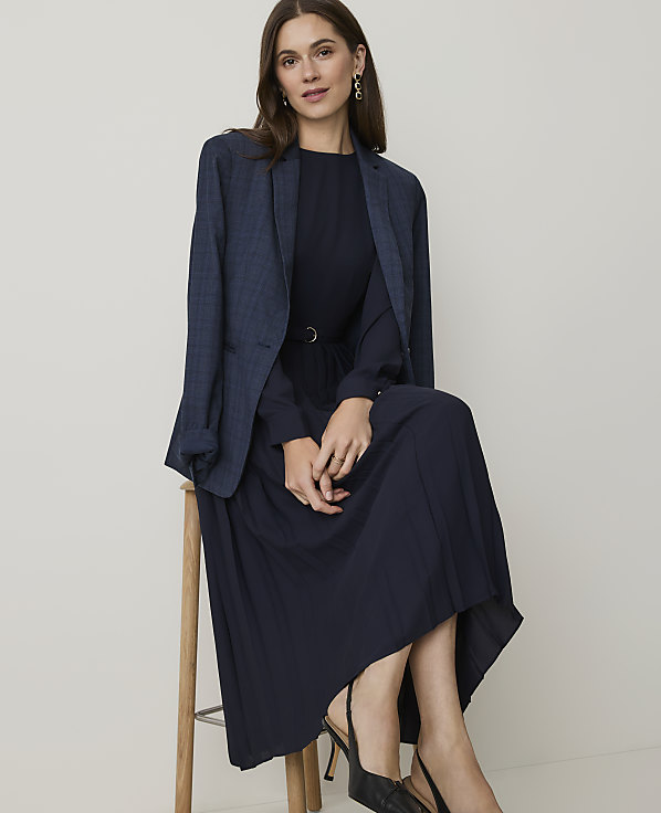 A woman in a navy dress and blazer sits on a stool against a plain white background.