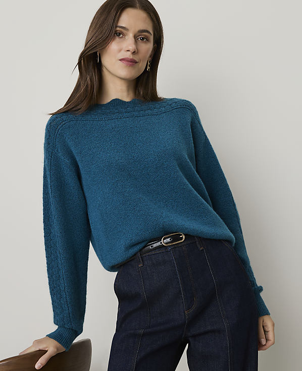 A woman in a teal sweater and dark jeans leans against a chair in front of a plain background.