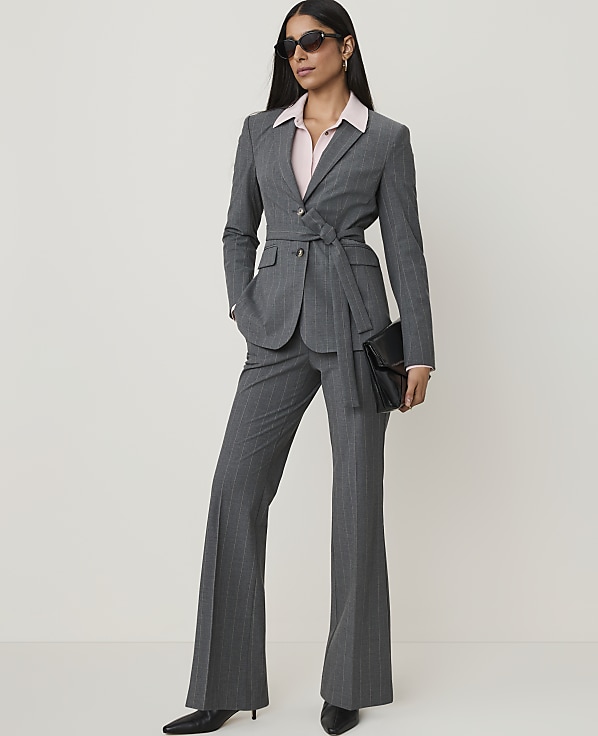 A woman in a pinstripe suit and sunglasses stands with a clutch in her hand against a plain background.
