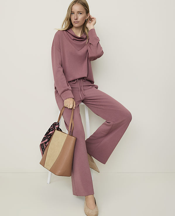 A woman in a mauve loungewear set and nude flats sits on a stool holding a tan and straw tote bag, against a plain, light-colored background.