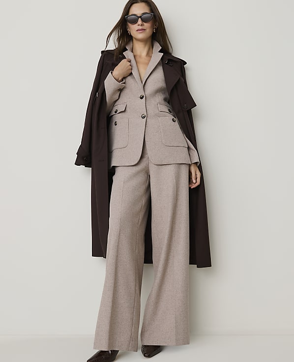 A woman in a beige suit and brown trench coat stands against a plain white background.