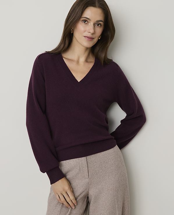 A woman in a plum v-neck sweater and beige pants stands against a plain gray background.