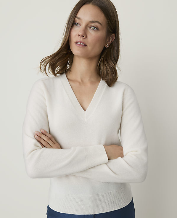 A woman in a cream v-neck sweater stands with her arms crossed against a plain, light background.